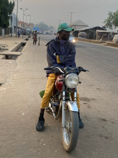 An outdoor, eye-level shot in Basawa, Nigeria, shows a man seated on a stationary motorcycle on a paved road under a hazy sky. The man, positioned centrally, wears a light blue or grey long-sleeved jacket, yellow trousers, a green baseball cap, and dark sandals. His expression is neutral, and he is looking towards the left side of the frame. He has dark skin and appears to be in his 30s or 40s.

The motorcycle is an older model with a prominent round headlight, a red or maroon fuel tank, a chrome front fender, and spoked wheels. The man's hands are on the handlebars. To the immediate left of the motorcycle, a dark object (possibly a bag or clothing) is draped over the seat.

In the background, the road stretches into the distance, with several other people visible further down the street, some walking. There are small, somewhat dilapidated buildings lining the road, consistent with a town or semi-urban area. Power poles with overhead lines are visible on the left. A concrete drainage ditch runs alongside the road, containing some debris. In the far distance, a vehicle's headlights are visible, suggesting either early morning or late afternoon/dusk, compounded by the soft, diffused light and hazy atmospheric conditions. No discernible text is visible in the image.