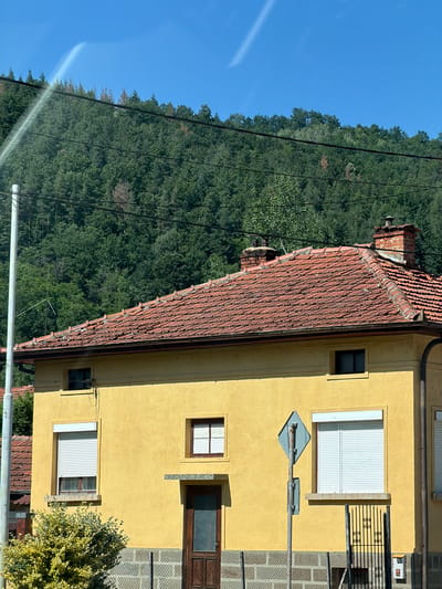 The image shows a yellow house with a red tile roof. The house has several windows, some with white shutters, and a wooden door. A brick chimney is visible on the roof. Behind the house, a dense forest covers a hillside under a clear blue sky. Power lines are strung across the scene. The location is identified as Beli Osam, Bulgaria. There are no people visible in the image.