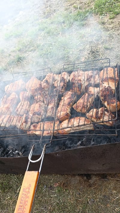 The image shows a close-up of a barbecue grill filled with numerous pieces of meat, likely pork ribs, cooking over hot coals. Smoke rises from the grill, obscuring some of the meat and creating a hazy atmosphere. The meat is charred in places, indicating it is well-cooked or nearing completion. The grill is positioned within a dark, metal brazier. A portion of the orange handle of a barbecue tool is visible in the lower left corner, featuring some lettering. The background consists of dry grass and soil, suggesting an outdoor setting, possibly a park or backyard. The image does not provide any specific clues about the location within the city of Паренда, Russia.