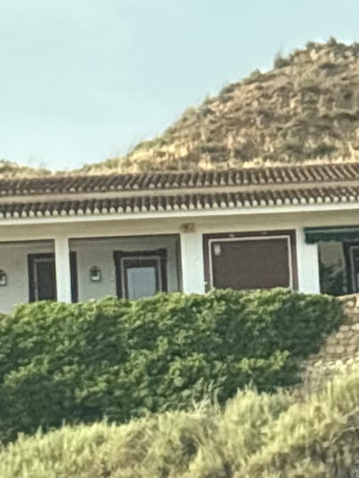 This is a vertically oriented, eye-level, medium shot of a building with an ornate facade, partially obscured by lush green trees. The building appears to be a mausoleum or columbarium, with a series of rectangular niches set into the wall. Each niche has a dark, recessed interior and a decorative frame. Some of the niches appear to be empty, while others contain what could be urns or other memorial items. The facade itself is light-colored, possibly stucco or stone, and features decorative trim along the edges of the niches. To the left of the building, a textured wall with what looks like a repeating patterned design is visible. To the right of the building, the dense foliage of a tree or shrub casts shadows on the facade and fills a significant portion of the frame. The scene is illuminated by natural daylight, suggesting it is daytime. The sky, visible in the upper left portion of the image, is a pale blue, indicating fair weather. The overall impression is one of a serene and solemn memorial site, possibly in a garden or cemetery setting. The architecture suggests a historical or traditional style. There is no visible text in the image.