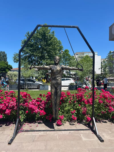 A bronze sculpture of a male gymnast is positioned outdoors. The sculpture depicts the athlete in a dynamic pose, holding gymnastic rings, with arms outstretched and legs slightly bent. It stands within a black metal frame that resembles an Olympic rings apparatus. In front of the sculpture, a dense bed of vibrant pink roses blooms, with some petals scattered on the paved ground. Behind the flowers, several cars, including a gray sedan and a white car, are parked. A light-colored scooter is also visible. In the background, there are trees, modern buildings, and a clear blue sky. A few people are visible in the distance, some appearing to be spectators or passersby. This sculpture is located in Sancaktepe, Turkey.