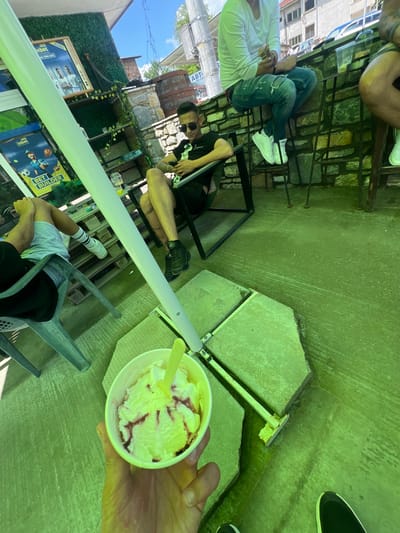 This detailed image captures an outdoor establishment, likely a casual cafe or a sports betting shop, situated in Касапите, Bulgaria. The scene is characterized by a pervasive green tint, potentially from an overhead canopy or reflective surfaces.

In the immediate foreground, a hand holds a white paper cup containing vanilla ice cream swirled with a reddish, possibly berry, sauce, and a yellow plastic spoon. A black shoe with a white sole is visible at the bottom right corner, belonging to the person holding the ice cream.

The mid-ground features several individuals. To the right, a man with dark hair, sunglasses, and visible tattoos on his left arm and leg is seated in a black metal chair. He wears a black t-shirt with a white design and black shorts, and is focused on a mobile phone in his hands. A clear plastic cup with a yellowish drink is on a nearby table. To the left, partially obscured, another person is seated in a grey plastic chair, wearing a light-colored t-shirt, shorts, and white socks with black stripes. Stacked wooden pallets are visible behind this person.

Further in the background, to the left, a man is seated on a low stone wall or barrier. He wears a white long-sleeved shirt, ripped blue jeans, and white Adidas sneakers, appearing to be engaged with an object in his hands. A bare leg and white sneaker of a fifth individual are visible on the far right.

The establishment's decor includes an artificial green wall serving as a backdrop for two "inbet" sports betting posters written in Cyrillic script. The upper poster advertises "НОВО" (NEW) and displays three men in sports jerseys, while the lower one promotes a "BET BUILDER" feature with illustrations of tennis, football, and American football. Two large wooden barrels and natural stone masonry further define the background. A prominent light-colored vertical pole stands in the center-midground, likely supporting the overhead structure.

The ground is a textured, light green concrete surface interspersed with paving slabs. In the distant background, a residential building with windows and a tiled roof is visible, along with parked cars on a street. A partially legible Cyrillic sign beginning with "АВТ..." and "НА С..." can be seen on a structure in the background. The overall scene depicts a relaxed daytime gathering.