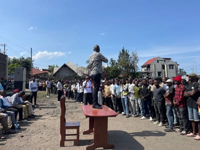 The image shows a group of people standing in a dirt lot. The location is in the city of Goma, Democratic Republic of Congo. A man wearing a camouflage-patterned shirt stands on a wooden table speaking to a group of people, approximately 60 men, mostly in their twenties and thirties. They are all listening to him intently. Some are wearing baseball caps. There is a single wooden chair on the ground in front of the table. There are two large speakers in the background, and other people are standing and listening to the speaker. There are buildings in the background. The weather is sunny and clear, and the lot is covered in dirt and gravel. Several men are sitting on the ground in the foreground. There are a couple of men with young children present. The image was likely taken during a public meeting, rally, or event.