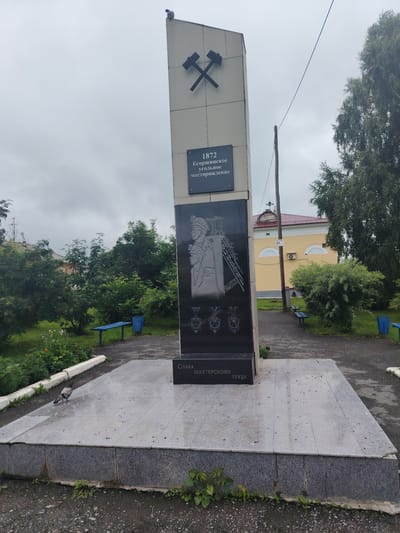 The image shows a monument in the city of Bulanasch, Russia, dedicated to the coal mining industry. The monument is a tall, slanted obelisk with a pair of crossed pickaxes at the top. Below the pickaxes is a dark plaque with text and a relief carving. The text reads "1872 Егоршинское угольное месторождение" which translates to "1872 Egorshinskoye coal deposit". The relief carving depicts three figures in miner's helmets and work clothes. Below the carving are three star-shaped emblems. At the base of the monument, another plaque reads "СЛАВА ШАХТЁРСКОМУ ТРУДУ" which translates to "Glory to the miner's labor". The monument stands on a granite platform. In the foreground, a pigeon walks on the platform. The monument is surrounded by greenery, including trees and bushes, and in the background, a yellow building with a red roof is visible. Two blue park benches and a blue trash bin are also present in the scene. The sky is overcast.