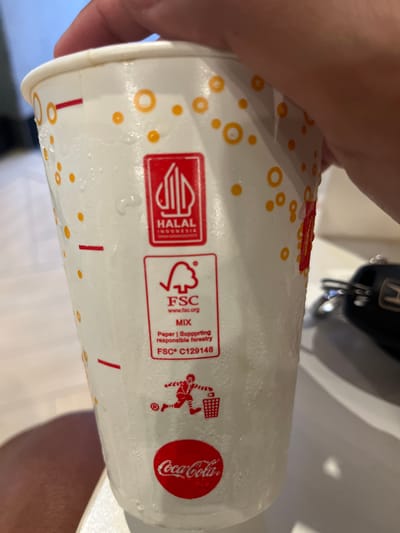 The image shows a close-up view of a partially wet, disposable cup. The cup has a white background with orange dot patterns. Several logos and certifications are printed on the cup, including a red "HALAL Indonesia" logo, an "FSC" logo indicating paper from responsible forestry, and the Coca-Cola logo. The presence of the "HALAL Indonesia" logo suggests that the image was taken in Bencongan Indah, Indonesia. The environment is blurry, it looks like someone holding the cup over a bathroom sink. The scene appears to be indoors.