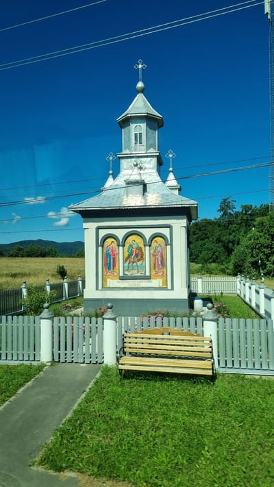 This is a full, eye-level shot of a small, white, Orthodox chapel with a silver, multi-tiered roof and a steeple topped with a cross. The chapel is decorated with three arched mosaics: one on the left depicting an angel, the center one showing a figure on horseback, and the right mosaic showing another angel. Below the mosaics are lines of text in Cyrillic. A grey, picket fence surrounds the chapel, with a paved walkway leading to its entrance. To the right of the walkway, there is a wooden bench on a patch of green grass. The chapel is set against a backdrop of a bright blue sky, distant green hills, and a field of dry grass. Power lines are visible across the top of the image. The location is identified as Krasnoilsk, Ukraine.