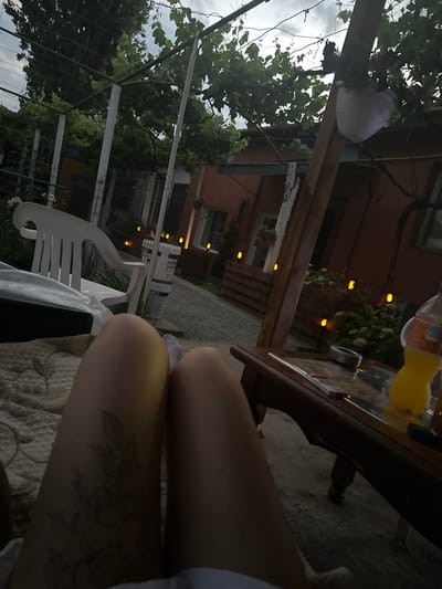 The image depicts an outdoor scene in the city of Golyam Chardak, Bulgaria. The foreground features a person's legs, with a subtle floral tattoo visible on the left thigh. They are resting on a beige patterned surface. In the middle ground, a white plastic chair is positioned to the left, and a wooden table with a glass top is on the right. On the table, there's an ashtray, some cards, and a partially visible yellow bottle of liquid. The background reveals a building with orange walls, a fence, and lush green foliage, including grapevines. Decorative orange lights are dotted throughout the scene, creating a warm ambiance. The sky appears overcast.