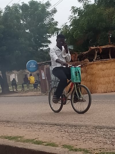 A man rides a bicycle on a paved road in Biliri, Nigeria. He is wearing a white button-down shirt, black pants, and sandals. He is holding a phone to his ear with his left hand as he rides. A blue bag hangs on the handlebars of the bicycle. Behind him to the left, a group of four people are sitting on a bench under a sign with the word "STAR" on it. The background includes trees, and buildings with thatched roofs.