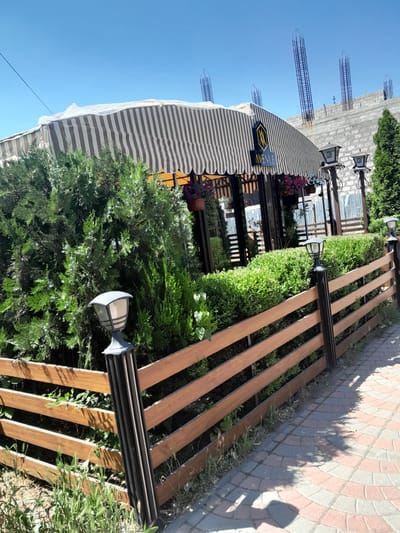 The image shows the exterior of a business with a striped awning. A wooden fence encloses an area with manicured hedges and trees. Several lampposts with black bases and grey tops are visible along the fence. A paved walkway with a patterned surface leads away from the fence. In the background, a building is under construction with rebar protruding from concrete structures. The sky is clear and blue. The scene appears to be a sunny day in Artashat, Armenia, possibly a restaurant or cafe with outdoor seating.