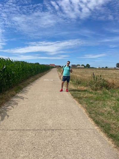 A man wearing a teal t-shirt, dark shorts, red sneakers, and a backpack stands on a concrete path in a rural area. The path is lined with a tall cornfield on the left and a dry, grassy field with scattered buildings in the distance on the right. The sky is blue with scattered white clouds. The image appears to be taken on a sunny day, likely in the summer. The location is the Kempen region of Belgium, near the village of Rooierheide in Diepenbeek.