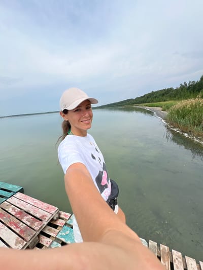 In Курорт Озеро, Russia, a woman wearing a white t-shirt and a light-colored baseball cap takes a selfie while standing on a wooden dock. The dock is constructed from colorful, weathered planks and is partially submerged in calm, greenish water. To the right of the woman, the shoreline is visible, featuring a sandy beach, tall reeds, and a dense forest in the background. The sky above is overcast with a pale blue hue. The woman is smiling and looking directly at the camera, with her right arm extended to capture the photo. She appears to be wearing a dark fanny pack across her chest.