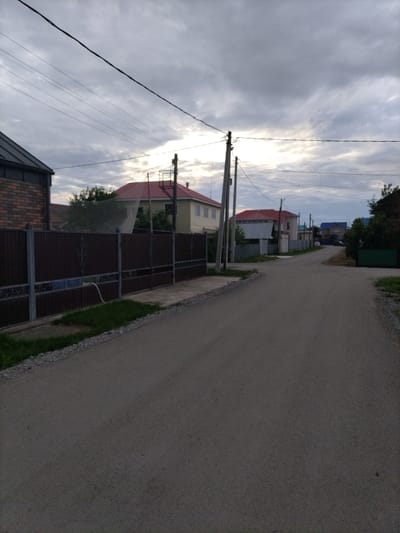 This media file depicts a quiet, rural street scene in the village of Nizhnyaya Makhtama, Russia, under an overcast sky. The road, paved with asphalt, stretches into the distance, curving slightly to the right. On the left side of the road, a tall, dark brown metal fence with decorative elements runs along the property line of a brick building with a dark roof. Beyond the fence, a light-colored house with a red roof is visible, partially obscured by trees and a white net. Further down the street on the left, more houses with red roofs and lighter facades are present.
On the right side of the street, there are also houses visible, one with a dark green gate and another with a blue roof further in the distance. The sky is filled with heavy, gray clouds, suggesting a cloudy or possibly impending rain. The presence of power lines and utility poles indicates that this is a residential area with access to electricity. The scene appears to be a typical day in a Russian village, with no active events taking place. The overall mood is peaceful and somewhat subdued due to the weather. The time of day is likely late afternoon or early evening, given the diffused light. No text is visible in the image.
