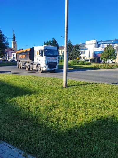 A white DAF semi-truck with an orange dump trailer is driving on a paved road. The truck has "XFORCE" written in orange on the side of the trailer and on the front of the cab. A number "95" is also visible on the side of the cab. The truck is in motion, and its tires are on the asphalt.
The scene is set on a sunny day with a clear blue sky. In the background, there are buildings with some trees and greenery. One building has "Top Diamonds" and "NABYTEK" written on its facade, indicating a commercial area. A church steeple is visible to the left of the frame, suggesting a town or city setting. The foreground is dominated by a lush green lawn, with a cobblestone path bordering the edge. The lighting indicates it is likely late afternoon, with shadows cast across the grass. There are no people clearly visible in the foreground. The overall mood is peaceful and bright.