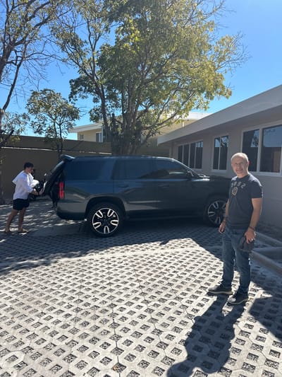 A rotated shot of a dark SUV parked on a patterned cement surface. An older man stands near the front passenger side of the vehicle, while a younger man stands behind it near an open door. Buildings are visible behind the car, with a blue sky and a tree branch framing the top of the image. The picture is somewhat underexposed, casting strong shadows on the ground.