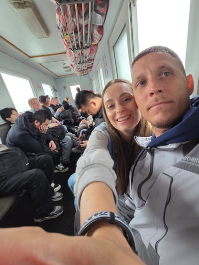 The image is a selfie taken from a tilted perspective inside a passenger transport vessel, likely a ferry or riverboat, characteristic of travel in a border region like Nizhneleninskoe, Russia.

In the foreground, a man and a woman are captured smiling towards the camera. The man, positioned on the left, has short dark hair, light stubble, and blue eyes. He wears a dark blue hooded top under a grey and white jacket, on which partially visible text reads "WORLD ORIGINATED IN 199[?]". To his right, the woman has long brown hair and a broad, joyful smile, also with blue eyes. She wears a light-colored long-sleeved top with a blue denim vest or jacket over it. A black and white patterned smartwatch is visible on her left wrist.

Behind the couple, numerous other passengers are seated in rows, extending into the background. Most passengers are seen from behind or profile, engaged in individual activities such as looking at their mobile phones or resting. The interior of the vessel features light-colored walls, large windows along the side that allow ample, diffused daylight into the cabin, suggesting it is daytime. Overhead storage racks are visible, filled with red life vests, each sealed in clear plastic for safety. The setting indicates a journey, with the couple appearing relaxed and happy, while other passengers seem calm and settled. The mix of passengers, including some of East Asian descent, further supports the context of cross-border travel from Nizhneleninskoe, Russia.