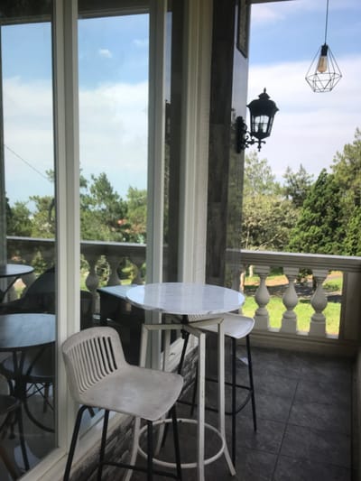 The image displays an unoccupied outdoor balcony or terrace area, likely belonging to a cafe or restaurant, situated in the city of Kota Batu, Indonesia. The scene is brightly lit by natural sunlight under a blue sky with scattered white clouds. In the foreground, a light-colored, possibly grey, bar stool with a slatted back and black metal legs is positioned. To its right, a round, high-top table with a white marble-patterned surface and a white metal frame stands, accompanied by a partially visible second white bar stool. The balcony floor is composed of dark grey tiles.
A white balustrade railing defines the perimeter of the balcony, overlooking a landscape of dense green trees, including conifers, and a strip of green grass visible below the railing. A dark-colored, possibly stone-clad, pillar supports a black, ornate wall-mounted lantern with a visible light source. Suspended overhead is a modern, geometric black wire-frame pendant light containing an incandescent bulb. The left side of the frame is dominated by large glass panels or sliding doors, reflecting the outdoor environment and an obscured interior or adjacent area containing additional tables and chairs. No individuals are present, and no actions or interactions are depicted.