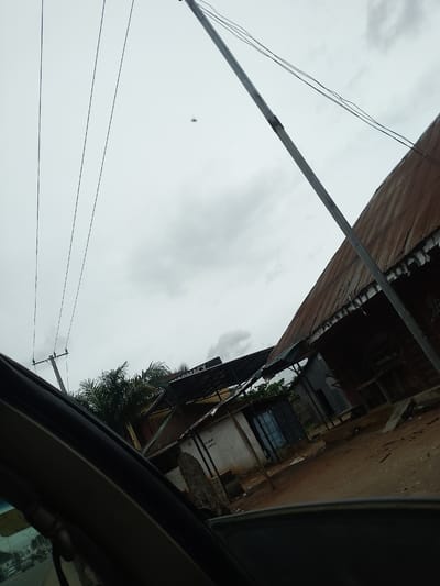 The image shows a street scene in Ubomiri, Nigeria, under an overcast sky. A power line runs diagonally across the frame, supported by a pole on the left. To the right, a building with a rusty corrugated metal roof is partially visible, with what appears to be a shop or business on the ground floor. Palm trees are visible behind the building. The foreground shows the interior of a vehicle, with the dashboard and window frame obscuring the lower part of the scene. The ground appears to be dirt or gravel. There are no people clearly visible in the image.