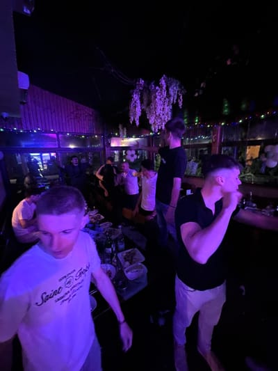 The image depicts a group of people at an outdoor gathering, likely a party, in what appears to be Panagyurski kolonii, Bulgaria, given the specific architectural features and the general atmosphere. The scene is lit with a combination of ambient light and string lights, casting a purple hue over the area.
Several individuals are visible: one man is lying down while other people are standing and interacting near tables with food and drinks. The table is cluttered with plates, food remnants, and bottles, suggesting ongoing socializing and dining. A glass-enclosed structure or greenhouse-like enclosure is present in the background, adding to the ambiance of the gathering. The scene conveys a sense of casual socializing and evening entertainment.