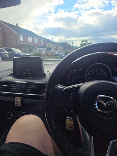 The image is a first-person perspective from inside a car, showing a view of a residential street in Lichfield, United Kingdom. The sky is partly cloudy, with bright sunlight breaking through. The car's dashboard is visible, including the steering wheel, air vents, and a central infotainment screen displaying a media player. A small religious icon is attached to the dashboard. Outside the car, a row of brick houses with pitched roofs lines the street, and several parked cars are visible. The driver's leg and knee are in the lower-left portion of the frame, suggesting the photograph was taken by the driver. The speedometer shows a speed of approximately 20 mph.