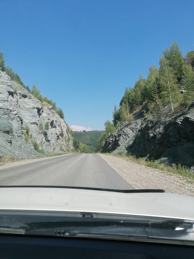 A POV shot from a vehicle traveling on a paved road through a mountainous landscape under a clear blue sky. The road curves gently to the right. On the left, a steep, rocky cliff face rises, sparsely covered with vegetation. On the right, a similar, though less steep, rocky incline is covered with trees, predominantly deciduous with some evergreens. The background shows rolling, green hills. The foreground shows the interior of the vehicle, with the dashboard and windshield wipers visible. The location is identified as the city of Severnoe, Kazakhstan.