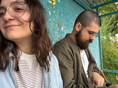Two individuals, a woman and a man, are seated side-by-side outdoors, likely at a bus stop or similar shelter, given the turquoise weathered wall and corrugated metal roof visible. The woman is in the foreground, closer to the camera, with her head turned to the left. She has dark, wavy hair and is wearing round-rimmed glasses and a light blue denim jacket over a striped shirt. Her expression is a slight smile. The man is positioned to her right, facing forward and looking down and to his right. He has short, dark hair, a beard, and is wearing glasses, a green jacket over a white t-shirt, and several bracelets on his left wrist. His hand, also on his left side, rests in his lap and has rings on his fingers. A dark umbrella is visible behind him. The background consists of the textured turquoise wall, with peeling paint and yellowed paper fragments. Beyond the shelter, greenery and trees are visible. The overall scene suggests a casual, everyday moment. The location is identified as Nikulskoye, Russia.