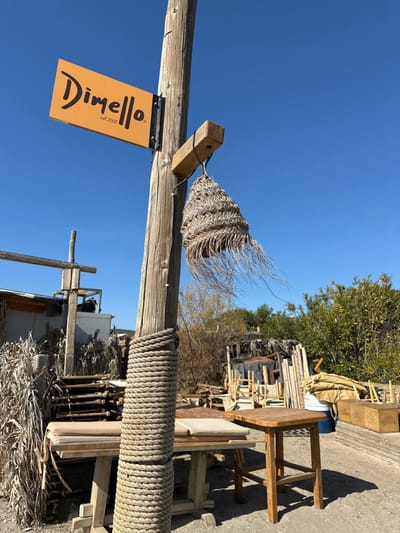 The image captures a rustic outdoor scene under a clear, bright blue sky, indicative of a sunny day, likely in Anatoliki Attiki, Greece. A prominent, weathered wooden post stands vertically on the left side of the frame.

Attached to this post is a rectangular, light-brown sign bearing the text "Dimello est. 2002". "Dimello" is written in a dark, stylized script font, with "est. 2002" in smaller, dark, sans-serif letters below it. The sign is secured with dark metal brackets.

Above the sign, a short, horizontal wooden beam is affixed to the main post. From this beam hangs a decorative, conical object fashioned from woven, light-colored, natural fiber, resembling straw or dried palm leaves. Its ends are frayed, giving it a windswept, organic appearance. Further down the main post, a thick, light-colored rope is tightly coiled multiple times around it, adding to the rustic aesthetic.

The background reveals an outdoor area with various wooden structures and elements. To the right, there's an assortment of wooden furniture, including a simple table and a stool, alongside stacks of what appears to be lumber or other construction materials. Green foliage from trees is visible in the upper right portion of the background. The ground seems to be unpaved, possibly dirt or gravel. There are no people or specific activities depicted. The scene suggests an outdoor establishment or a storage area for natural, handcrafted elements, embodying a casual and natural ambiance.