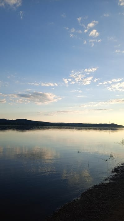 The image depicts a tranquil landscape at dusk or dawn in Шолья, Russia. The scene is dominated by a wide body of calm water reflecting the sky, with a distant treeline and a pebbly shoreline in the foreground.
The sky occupies the upper two-fifths of the frame, presenting a gradient from medium blue in the upper left to a pale yellow-orange hue on the horizon to the right. Scattered white cumulus clouds are dispersed, more densely clustered in the lower half of the sky. Several small, dark specks, likely birds, are visible in flight against the blue sky, particularly in the upper right quadrant.
A dark, undulating horizon line, consisting of a forested landmass or hills, stretches across the mid-ground, separating the sky from the water. The silhouette of dense tree cover is visible. Towards the far right of this distant land, faint structures resembling industrial pylons are discernible against the lighter sky.
The water surface is smooth, exhibiting clear reflections of the sky and clouds. The reflection directly below the sky maintains a similar blue and pale yellow coloration, with the white clouds distinctly mirrored. The water closer to the foreground appears darker blue, with subtle ripples evident, particularly near the bottom right. A few slender reeds or blades of grass emerge from the water along the right edge.
The immediate foreground, in the lower right corner, consists of a dark, coarse shoreline composed of small pebbles or gravel, gently sloping into the water. No individuals or specific human-related actions are depicted.