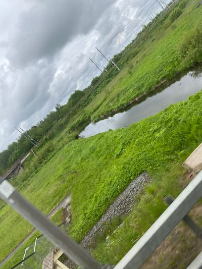 The image presents a view from an elevated position, possibly a train or bridge, overlooking a canal or narrow river. The location is assumed to be near Kommunar, Russia due to the presence of infrastructure typical of that area. The water body is flanked by dense green vegetation, predominantly grass and bushes. Metal railings, likely part of the structure from which the image was taken, are visible in the foreground. The sky is overcast with heavy clouds. Power lines with supporting pylons run along the far side of the water feature. There are no people visible in the image. The overall impression is of a rural or semi-rural scene viewed from a railway or similar infrastructure.