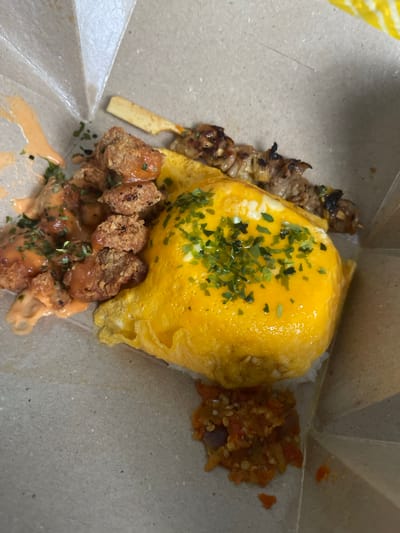 The image displays a meal served in a light brown cardboard takeout box, typical of street food or casual dining in a region like Ngasinan, Indonesia. The central and most prominent item is a large, fluffy yellow omelette, generously sprinkled with chopped green herbs, likely parsley or chives, resting on a bed of white rice.

To the left of the omelette, a wooden skewer holds several pieces of grilled meat, exhibiting char marks. Below the omelette, a portion of crispy fried chicken pieces is visible, coated in a creamy, orange-hued sauce and also garnished with green herbs. Completing the meal, a small pile of vibrant red and orange chili paste, known as sambal, with visible seeds and chopped ingredients, is placed to the right of the omelette. No text is visible in the image.