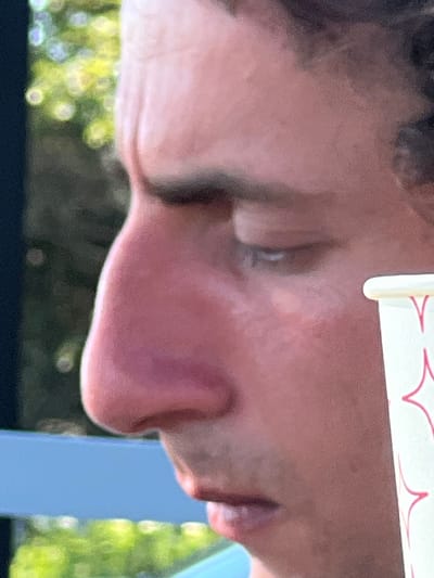 The media file is a close-up, side profile of a person's face, primarily featuring their nose, mouth, and chin. The image is oriented vertically, with the top of the person's head at the bottom of the frame and their feet at the top. The person appears to be young, with smooth skin and a light complexion. Their mouth is slightly open, and their lips are full. A portion of a white paper cup with red decorative lines is visible at the top of the frame, suggesting the person might be holding or drinking from it.

The background is blurred, but hints of green foliage and a blue object are discernible, implying an outdoor setting. The lighting suggests it could be daytime, with soft light illuminating the person's face. The overall impression is one of casualness, possibly a candid moment.

No specific activity or event is clearly depicted, other than the person's relaxed posture and presence with a beverage. The emotional state is neutral or relaxed, with no strong indicators of happiness, sadness, or distress.

There is no discernible text in the image, other than the abstract red design on the paper cup. The location context of Ochamchire, Abkhazia, does not provide any direct visual cues within the frame itself.