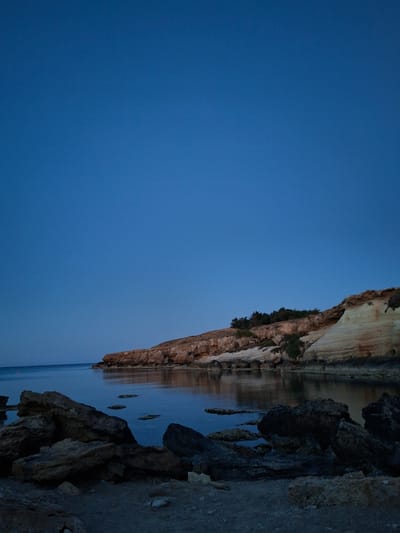 The image displays a tranquil coastal landscape, likely at twilight or dawn, given the deep blue hues of the sky and water and the soft, indirect lighting. A prominent, layered cliff face with shades of tan and brown rock, dotted with dark green vegetation, occupies the right side of the frame. This cliff is clearly reflected in the calm, dark blue water that extends across the lower and central parts of the image. In the foreground on the right, dark, rugged rock formations and a narrow stretch of shore provide visual depth. Above the distant cliff and water, the sky is a vast, clear expanse of deep blue. There are no visible people or activities, emphasizing the natural serenity of the scene. No text is visible. The tranquil setting is characteristic of a coastal area in Paralimni, Cyprus.