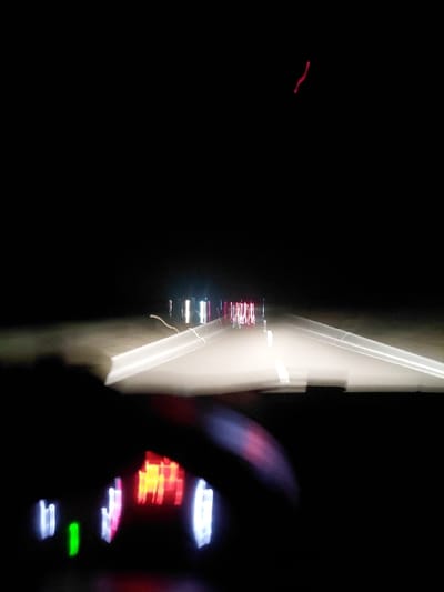 A blurry, long-exposure shot captures a nighttime scene from inside a moving vehicle. A dashboard with illuminated gauges glows on the right. The road ahead is visible as a bright streak of light with faint hints of oncoming headlights and taillights. The left side of the image is predominantly dark, showing a faint red streak. The motion-blurred lights suggest the vehicle is traveling at a significant speed.