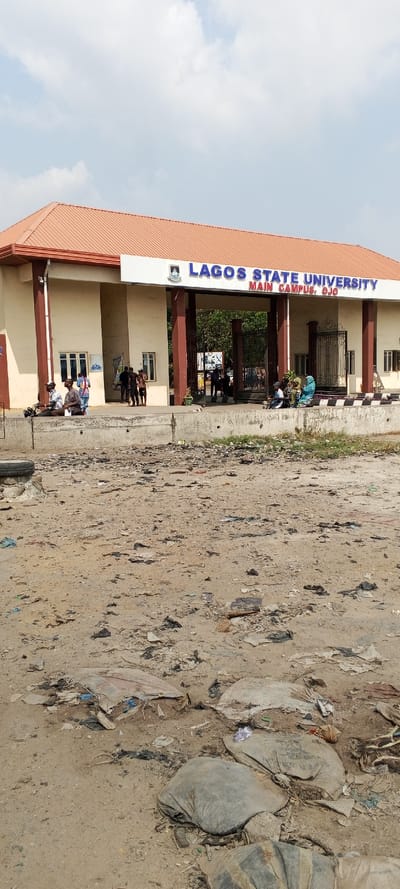 The image depicts the main entrance of Lagos State University, Main Campus, Ojo, situated in Awjaw, Nigeria, under a bright, partly cloudy sky. The foreground consists of an unpaved, sandy-dirt area heavily littered with various debris including plastic bags, paper, fabric scraps, and multiple discarded tires or sacks.
The midground features a prominent, single-story entrance structure with a reddish-brown hip roof and light beige walls supported by dark brown pillars. A large sign affixed to the front facade reads "LAGOS STATE UNIVERSITY MAIN CAMPUS, OJO" in blue lettering, accompanied by a university crest. Beneath the sign is a large, ornate black metal gate, partially open, providing access to the campus interior where additional foliage and structures are visible. Flanking the gate are two smaller structures with windows, likely guard posts or administrative offices. A concrete barrier with a checkered pattern on its top edge separates the unpaved foreground from the elevated entrance area.
Approximately ten to fifteen individuals are observed around the entrance. On the left side of the concrete barrier, two individuals are seated, one near a small motorcycle, with another person standing nearby. Near the main gate, a group of about five to six individuals stands and interacts. On the right side of the concrete barrier, at least three individuals are seated, one prominently draped in colorful fabric. Additional individuals are visible deeper within the gated campus area. The overall scene indicates an active university entry point with pedestrian presence.