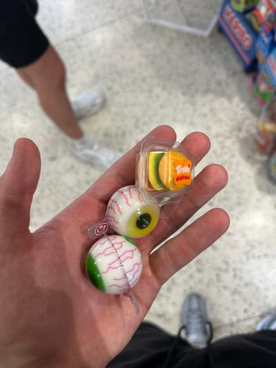 A person's hand holds two Trolli Eyeball candies and a small Trolli Originals hamburger candy. The candies are in clear plastic packaging. The hamburger candy has a yellow bun and a green patty. The eyeball candies are white with pink veins and green bottoms. In the background, other candies are visible on shelves. The person is wearing black pants and white sneakers. The setting appears to be a store.