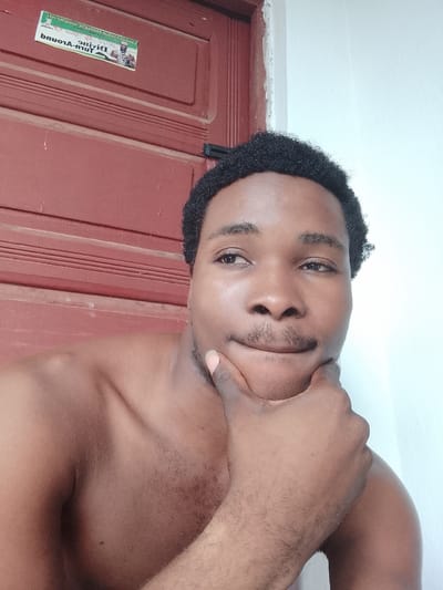 A close-up, waist-up, outdoor, natural-light, selfie photograph is captured in Orodo, Nigeria. The subject is a young Black male with dark, curly hair and a light mustache. He is bare-chested and resting his chin on his right hand, with his left hand on his chest. The background consists of a red wooden door with horizontal planks and a section of a light-colored wall. A sticker with text and an image of a man is affixed to the upper left portion of the door. The image is slightly tilted, with the subject's face angled towards the camera and his eyes looking slightly to his right. The lighting is soft, creating subtle shadows on the subject's skin and the background. The overall impression is one of a casual, personal moment captured in an everyday setting.