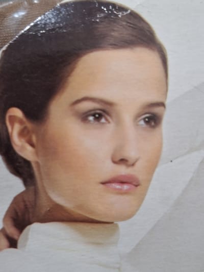 The image is a close-up portrait of a woman. She has brown hair pulled back, and her face is turned slightly to her left. Her eyes are brown and her lips are full and painted with a pale pink lipstick. She is wearing a white turtleneck. The background is out of focus and appears to be a light gray or white. There is no indication of the city of Ötegen batyr, Kazakhstan in the image.