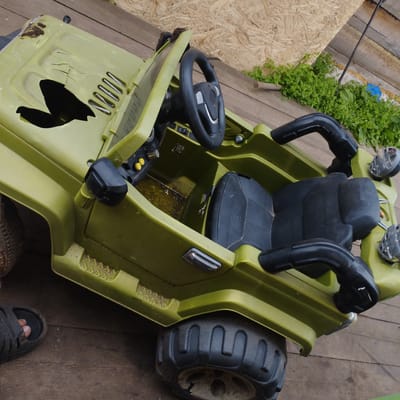 A damaged, olive-green toy jeep is situated outdoors on a wooden surface. The jeep exhibits significant wear and tear, including a large crack and hole in the hood, rust on the front, and a broken front windshield frame. Inside the jeep, a black seat is visible. The vehicle has large, rugged tires. In the background, a patch of green vegetation is present, along with some weathered wooden planks. A person's foot wearing a black sandal is visible on the lower left side of the image, suggesting the jeep is on the ground. The scene appears to be in a residential outdoor area, possibly a yard or alley, in the village of Pokosnoye, Russia.