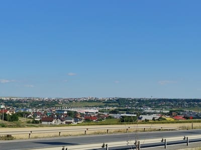 The image captures a wide-angle view of a suburban landscape on a clear, sunny day, likely taken from an elevated position overlooking a city or town. In the foreground, a section of a highway is visible, with a light-colored guardrail separating the road from a field of dry, yellow vegetation, possibly wheat or hay. The road itself is asphalt with white lane markings.

Beyond the highway and fields, a sprawling residential area is depicted. Numerous houses with red, brown, and gray roofs are clustered together, interspersed with trees and greenery. Some of these appear to be single-family homes, while others might be townhouses or low-rise apartment buildings. Further in the distance, a denser urban area unfolds, with taller buildings and a more commercial or industrial appearance, including what looks like a shopping center or industrial park with large, light-colored structures and some with red roofs. A water tower stands prominently against the skyline in the far distance. The overall impression is one of a developing urban fringe. The location is identified as Șcheia, Romania. There are no people or animals visible in the image.