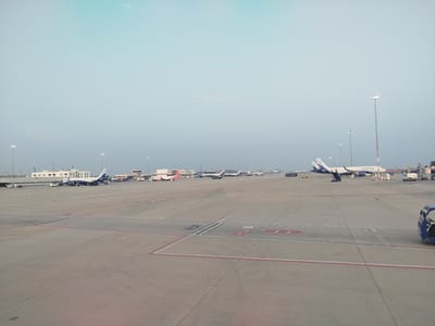 The image captures a wide-angle view of an airport tarmac under a clear, bright sky. Numerous airplanes are visible, predominantly featuring the white and blue livery of IndiGo Airlines, with "IndiGo" text clearly discernible on their fuselages. At least five to six IndiGo aircraft are parked or taxiing in the mid-ground and background. One aircraft in the left background appears to have orange accents, possibly belonging to another airline.
The tarmac, made of concrete, stretches across the foreground and mid-ground, featuring various red, white, and black markings, including numerical and directional indicators. Partially visible text on the ground includes "EAST". Tall light poles equipped with multiple lamps are strategically placed throughout the airport grounds, some appearing to be illuminated.
In the background, airport terminal buildings and what appears to be a control tower are visible, indicating a large and operational airport. Ground support equipment, such as mobile stairs, baggage carts, and other utility vehicles, can be seen near the parked aircraft. A distinctive blue ground vehicle is partially visible on the far right side of the frame. The sky is a light blue, transitioning to a slightly greenish-blue hue at the top. The lighting suggests a clear day, possibly early morning or late afternoon, with soft, ample light. The scene depicts a typical busy airport environment, consistent with the specified location in Anneshvara, India. Visible text includes "IndiGo" on planes, "60" on a small sign, and "TJD" on a ground vehicle.