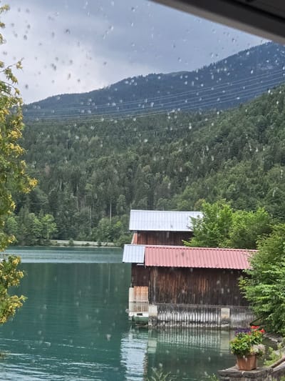 A serene, overcast day is captured through a window with water droplets, showing a picturesque view of Kochel am See, Germany. The foreground features a calm, teal-colored lake with subtle ripples. On the right side of the lake, two wooden boathouses with corrugated metal roofs are visible, one with a silver roof and the other with a reddish-pink roof. A potted plant with red and purple flowers sits on a stone ledge in front of the boathouses.

Behind the lake, a dense, green forest covers the slopes of a large mountain. Power lines are strung across the mountain face, partially obscured by the trees. The sky is a muted gray, indicating cloudy weather. A branch of a yellow-leafed tree is visible on the far left, adding a touch of color to the scene. The overall impression is one of peaceful, natural beauty, enhanced by the soft light and the presence of the lake and surrounding landscape.