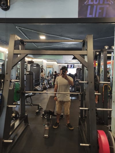 An adult male is observed taking a selfie in a gym, reflected in a large wall-mounted mirror. The image does not contain information to confirm its location in Staufenberg, Germany. The man, whose face is obscured by a smartphone, wears a light-colored t-shirt, patterned knee-length shorts, dark sneakers, a wristwatch on his left wrist, and a dark baseball cap worn backward. He stands centered in front of a dark gray metal squat rack or Smith machine, which has a barbell racked at approximately chest height. A black adjustable weight bench is positioned on the dark rubber flooring in front of the squat rack. Various weight plates, including a green and a red one, are visible on equipment or racks. In the background, other gym equipment such as weight machines and additional individuals are visible, indicating an active gym environment. Overhead lighting fixtures illuminate the space. A partial sign with purple text reading "LOVE LIFT" is visible on the upper right side, reflected above the gym equipment.