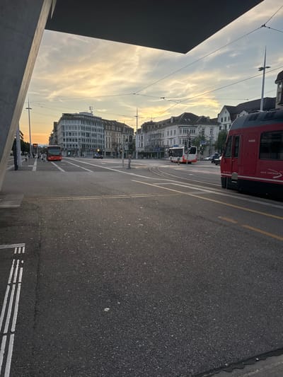 The image depicts an urban street scene at dusk in Solothurn, Switzerland, specifically at Bahnhofplatz, looking towards the city center from the vicinity of the main train station.
In the immediate foreground, a grey concrete structural element, likely part of a building or canopy, occupies the upper left, while the ground is dark grey asphalt pavement with white striped tactile paving and double yellow road lines.
The midground features a wide, multi-lane asphalt roadway with distinct white lane markings, pedestrian crossings, and a complex network of tram tracks, with a prominent curve to the right. Overhead power lines for public transport span across the scene. Traffic signals are visible at the intersection.
Multiple vehicles are present: A red and white public bus is positioned on the far left, near a sidewalk. A dark sedan is visible further down the street in the center. A white and orange public bus is located on the right side of the main intersection. Partially cropped on the far right, a red train or tram, marked "aare seeland mobil" and "VENUS," is stationary on the tracks.
Several indistinct figures, appearing to be pedestrians, are visible on the sidewalk to the left, near the red bus.
The background is defined by multi-story urban buildings of varying architectural styles, from modern grey structures on the left to older, ornate white and brown-roofed buildings on the right. Streetlights are also visible.
The sky is partially cloudy, displaying a blend of light blue and warm orange-yellow hues near the horizon, consistent with a low sun angle during dusk.