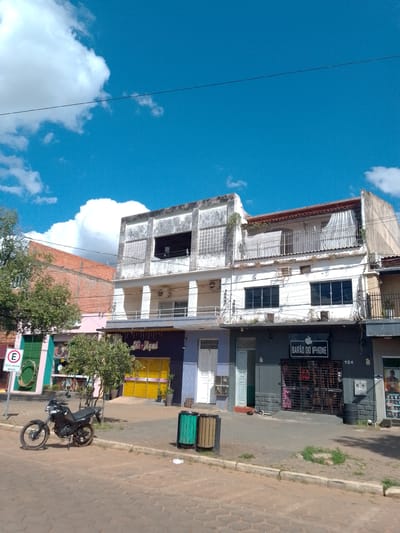 The image shows a street scene in Cáceres, Brazil. In the foreground, a black motorcycle is parked on a cobblestone street. To the right of the motorcycle are two trash bins, one green and one brown. Behind the motorcycle and trash bins, a row of buildings lines the street. These buildings appear to be commercial establishments, with storefronts and upper-story windows. One storefront has a sign that says "BARÃO DO IPHONE" with an Apple logo. The sky above is bright blue with scattered white clouds. Trees and vegetation are visible in front of the buildings. The overall impression is a sunny day in a Brazilian city.