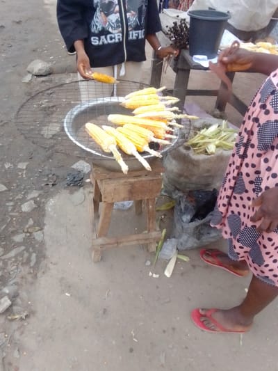The scene takes place on a dirt road in Sapele, Nigeria, at the coordinates 5.95 latitude and 5.695 longitude. A person wearing a black shirt with white lettering, possibly "Sacrific," and khaki pants is standing on the left side of the frame, holding a corn cob. A woman with a pink polka-dotted dress is standing on the right side of the frame. She has her right arm extended and appears to be gesturing. A pile of husks and corn cobs is in a woven bag on the ground. There is a round, metal bowl with a wire rack and a wooden stool, on which the bowl is resting. Inside the bowl is a charcoaled fire, with corn cobs, about 12 in total, on the rack. Most of the cobs are charred and partially cooked. Behind the bowl is a wooden table on which there is a black bucket. There are some other miscellaneous items on the table, some of which are out of frame. On the ground, the dirt road has pieces of gravel and stones, with scattered husks and other plant matter. The scene is partially lit, with shadows cast across the road. There is an uneven surface, indicating that the location is not paved. The people are interacting. It appears that the woman is a vendor and is selling the charred corn cobs. The man on the left side of the frame is potentially buying or just observing the process.