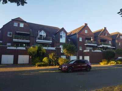 This image shows a street in Leverkusen, Germany, featuring a row of brick apartment buildings with pitched roofs and multiple balconies. A maroon Volkswagen Golf is parked on the right side of the street, facing left. The buildings have white garage doors at ground level. Trees and bushes are visible in front of the buildings and along the street. The sky is clear and blue, and the sunlight casts long shadows, indicating it is likely late afternoon or early morning. The overall scene depicts a residential street in a European city.