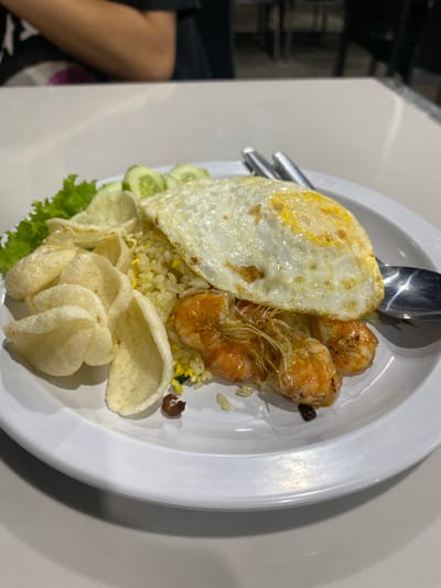 A close-up, eye-level shot shows a white plate filled with nasi goreng, or Indonesian fried rice, with a fried egg and several cooked shrimp on top. The rice is a light yellow color with visible specks of green and brown. The fried egg has a perfectly cooked yolk. The shrimp are orange and appear glazed. Scattered around the rice are several light-colored, puffed crackers, some lettuce leaves, and sliced cucumbers. A fork and spoon are resting on the plate. The plate is on a white table. In the background, out of focus, a person wearing a black t-shirt is seated at the table. The scene is likely a restaurant in Mekar Bakti, Indonesia, given the context of the query.