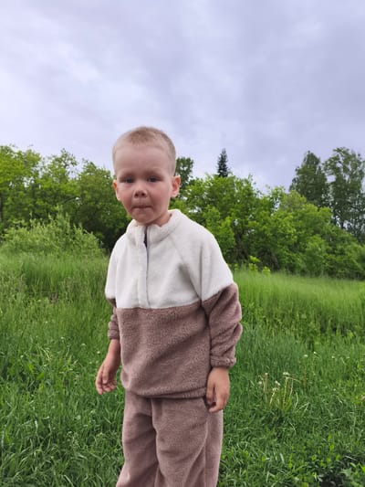 The image depicts a young child, approximately 2-4 years old, standing outdoors in a grassy field in the city of Можга, Russia. The child, with fair skin and short, light-colored hair, is positioned centrally in the frame, looking directly towards the camera with a neutral expression, exhibiting slightly pursed lips. The child is wearing a two-piece fuzzy fleece tracksuit: the upper portion of the top is off-white, while the lower portion of the top, sleeves, and pants are a uniform light brown or taupe. The top features a short zippered placket at the neckline.
The immediate foreground consists of dense, verdant green grass with scattered small white wildflowers and some dried plant stalks. Behind the child, a field of tall green grass and lush green foliage of various bushes and trees extends into the midground and background. A discernible line of deciduous trees forms the main treeline, with one taller, darker evergreen tree visible to the left within the tree line. The sky above is overcast and light grey, indicating cloudy weather. The overall setting is natural and rural, dominated by green vegetation under a muted sky.