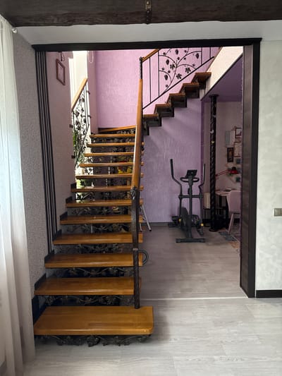 This media file presents an interior view of a multi-level home in Krapivka, Russia, focusing on a prominent staircase descending from an upper floor. The staircase features light-colored wooden treads and risers, complemented by a striking black wrought-iron railing adorned with intricate grape and leaf motifs, topped with a dark wooden handrail.
At the upper landing, a stationary exercise bike, primarily black and gray, is positioned on light wood-look flooring. The walls in this upper area are covered with a textured, crinkled purple wallpaper. Beyond the exercise bike, in an adjacent room, framed pictures are visible on the wall, along with a small red desk lamp and hints of furniture, possibly a chair or desk.
At the bottom of the visible stairs, a landing is covered with a light-colored carpet featuring a subtle patterned design. A dark wooden beam is visible on the far left, and another section of the ornate railing extends further down out of frame. Light wood-look flooring also appears on the right side of the lower area.
The scene is well-lit, suggesting daytime. No people are present, and there is no visible text. The overall aesthetic combines traditional elements, like the ornate staircase, with modern household items like the exercise equipment.