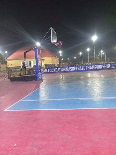 The image depicts a brightly lit outdoor basketball court at night. The court is painted in red and blue with white lines. A basketball hoop stands on the left side of the court. Behind the hoop is a yellow building with a red roof. A banner reads "DAM FOUNDATION BASKETBALL CHAMPIONSHIP" across the back of the court. Multiple light poles illuminate the scene.