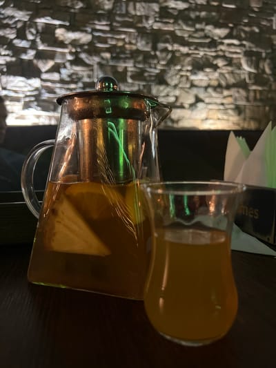 A glass pitcher with a built-in strainer and a glass cup are on a table, likely indoors. The pitcher contains a yellow liquid, possibly tea, with lemon slices visible. The cup is filled with the same liquid. The table is dark, and the background shows a textured stone wall. The location in the city of Zaosinovo, Russia is not discernible from the image.